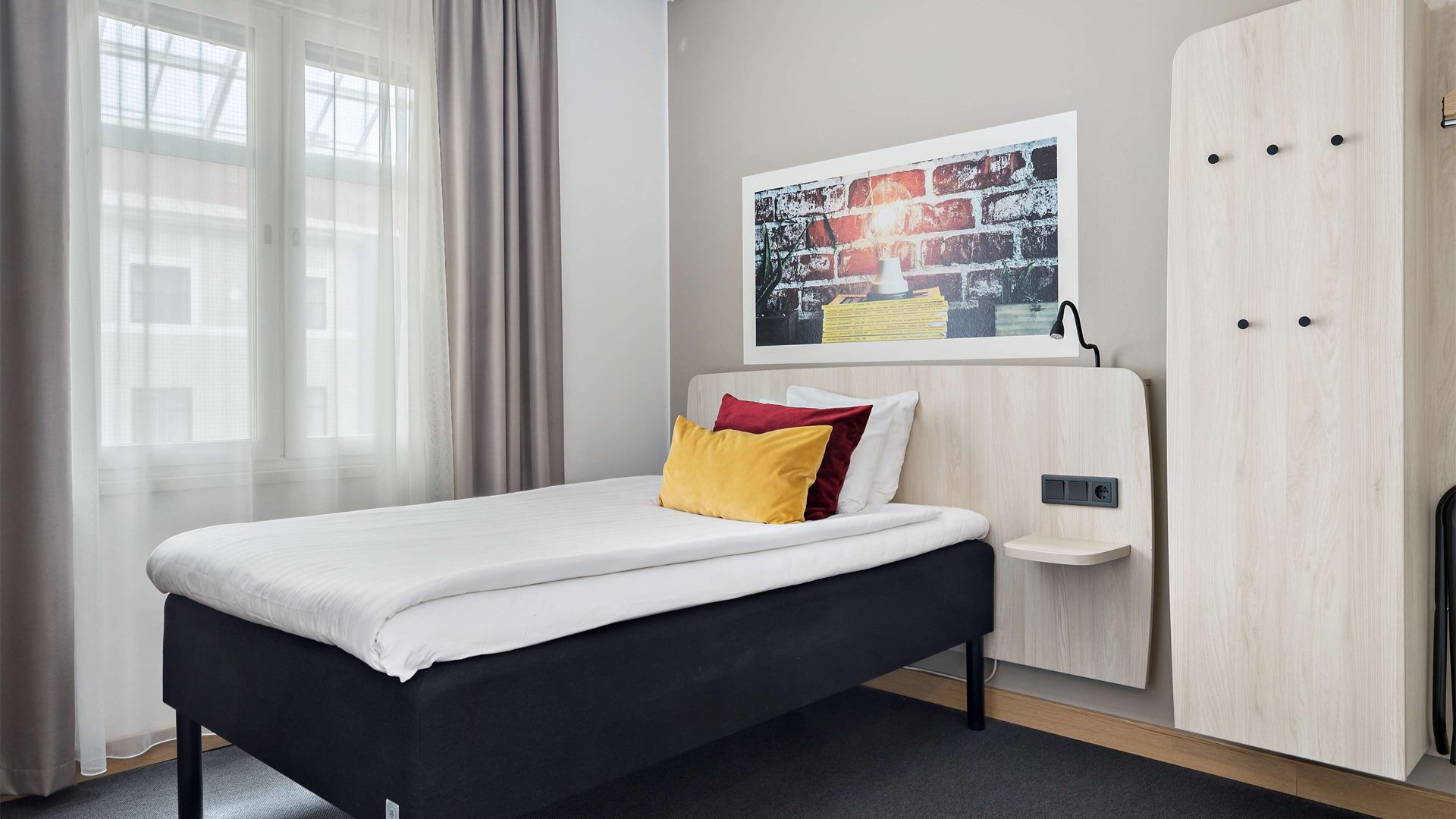 Ett lite mindre hotellrum med en enkelsäng. En tavla ovanför sänggaveln. Ett fönster släpper in ljus i rummet.