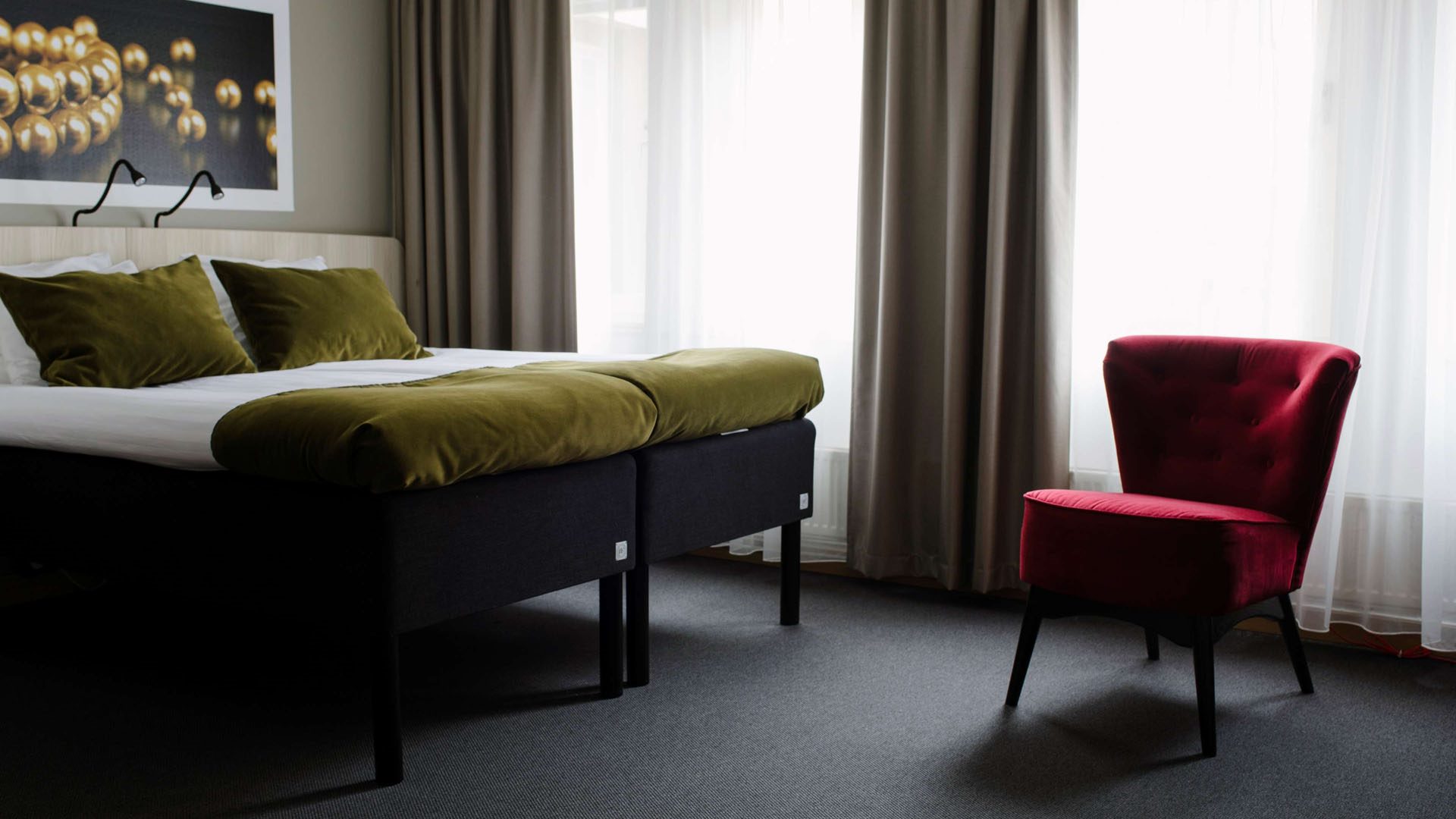 Hotellrum med en säng och en fåtölj. Över sängen hänger en tavla. Fönstren täcks av gardiner.