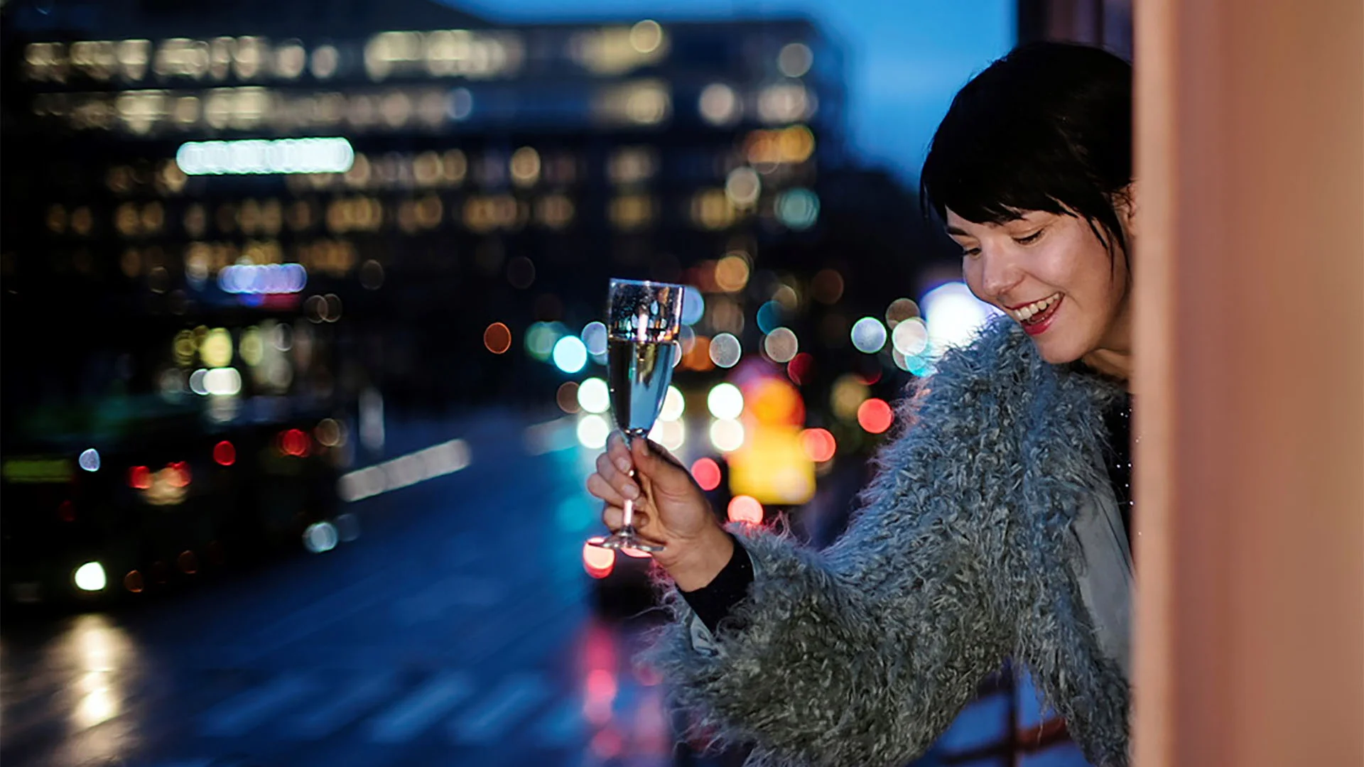 Kvinna hållandes champagneglas lutar sig ut från ett fönster. Stad i bakgrunden.