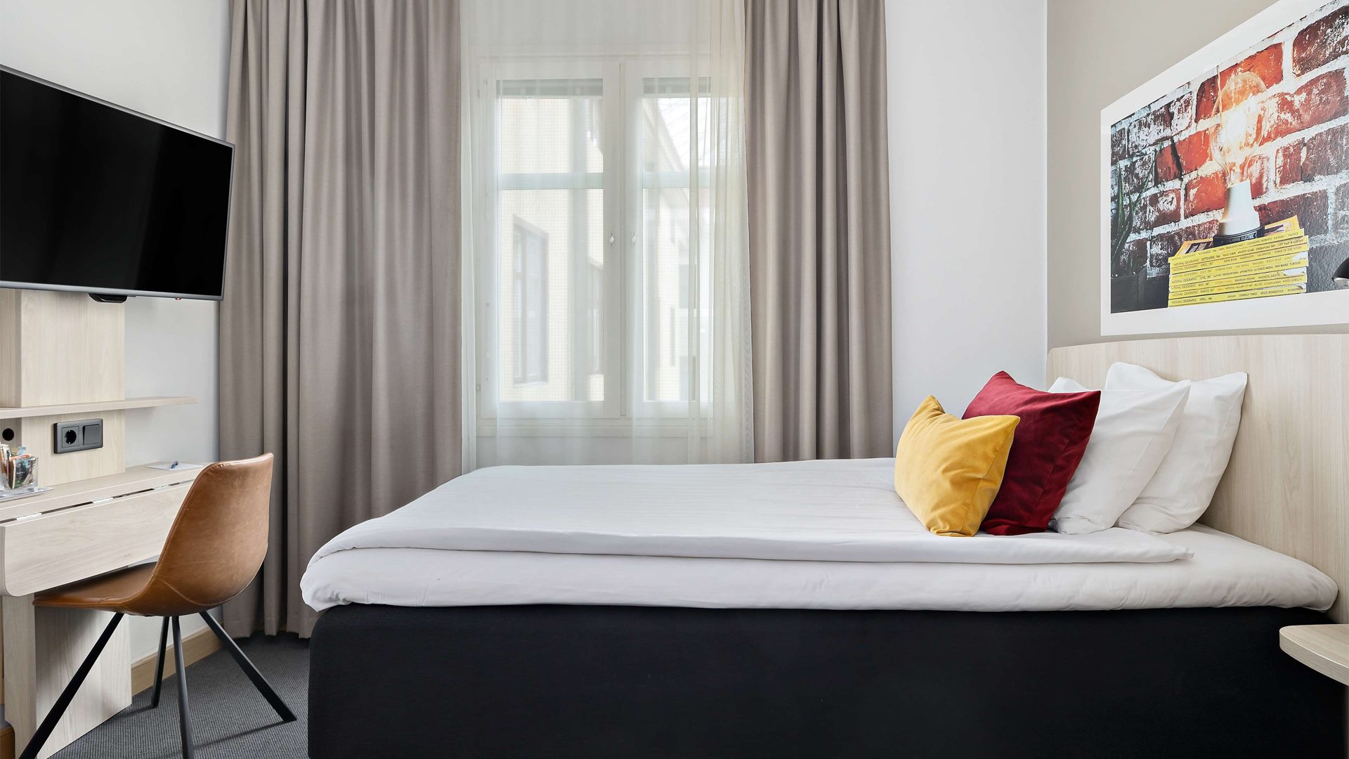 Hotellrum med säng, fönster och skrivbord. En tv hänger på väggen.